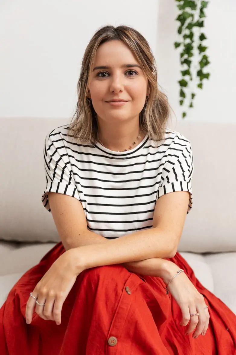 Bruna está sentada em um sofá, vestindo uma camiseta listrada de branco e preto e uma saia vermelha. Ela está sorrindo levemente e olhando diretamente para a câmera. Ao fundo, há uma planta pendente.