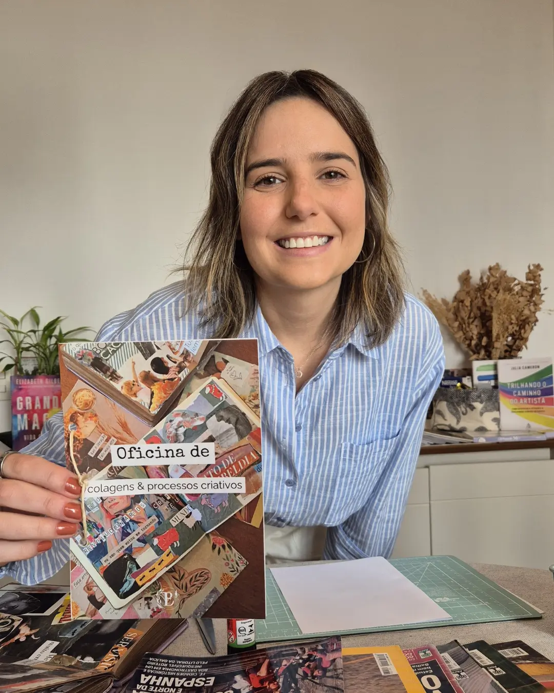 Bruna está sorrindo e olhando diretamente para a câmera, segurando o encarte da Oficina de Colagens e Processos Criativos.